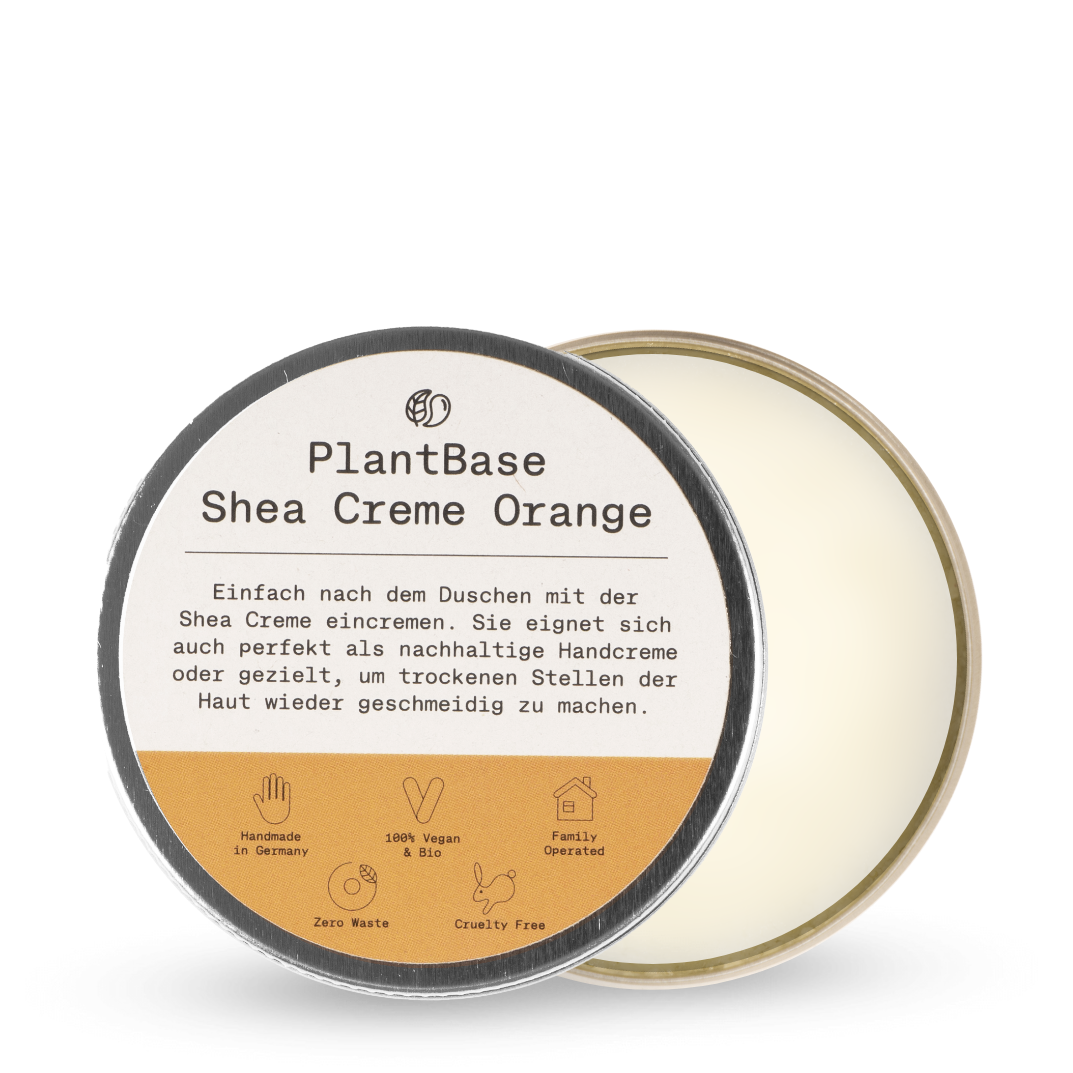 Eine runde Metalldose mit PlantBase Shea Creme Orange, bei der der Deckel teilweise geöffnet ist, so dass man die Creme im Inneren sehen kann. Das Etikett enthält Produktdetails auf Deutsch und Symbole für handgemacht, vegan, abfallfrei und tierversuchsfrei.