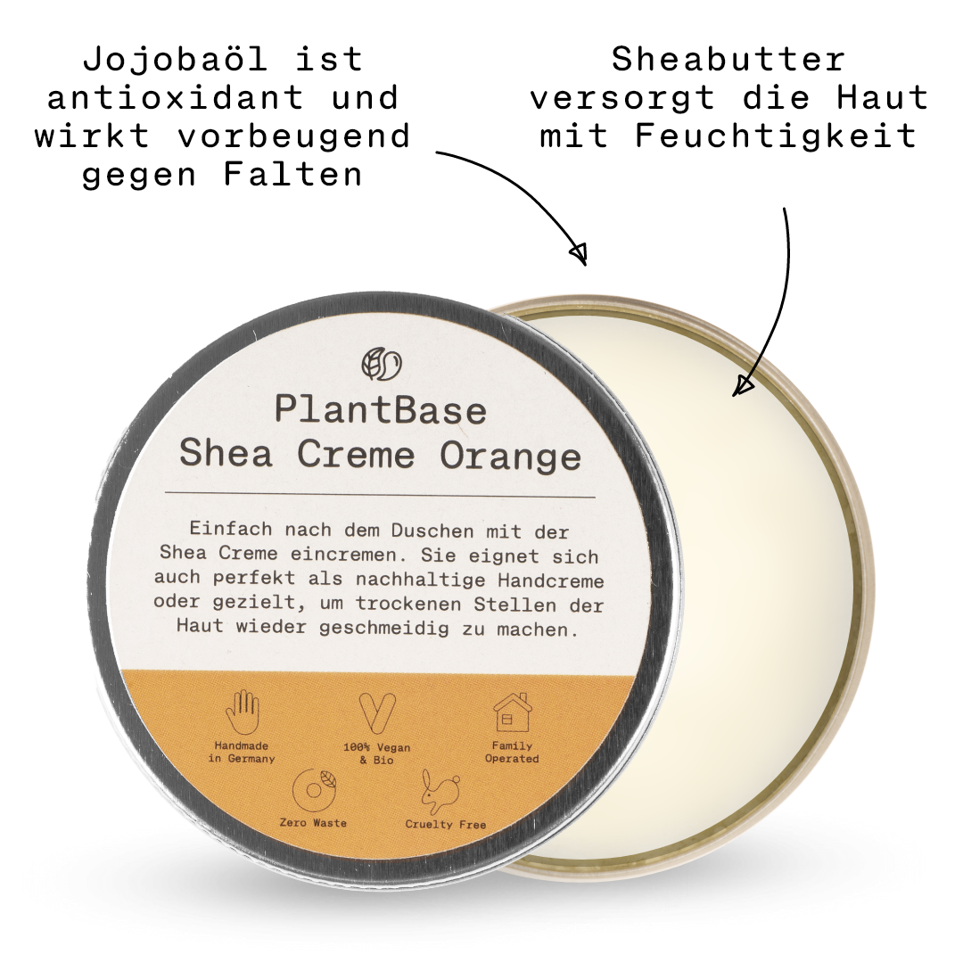 Eine runde Dose mit einem beige-weißen Etikett mit der Aufschrift "PlantBase Shea Creme Orange". Der geöffnete Deckel enthüllt das cremige Produkt im Inneren. Symbole heben hervor, dass es handgemacht, vegan, tierversuchsfrei und abfallfrei ist.