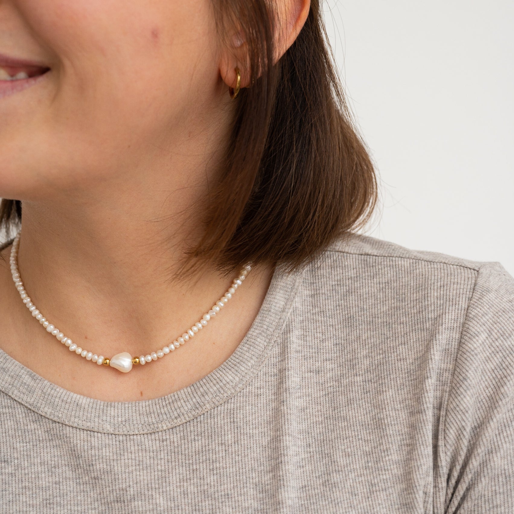 Eine Frau trägt ein hellgraues geripptes T-Shirt und eine Perlenhalskette mit einer großen Perle in der Mitte. Ihr kurzes braunes Haar hat sie hinters Ohr gesteckt, und sie trägt einen kleinen goldenen Ohrring.