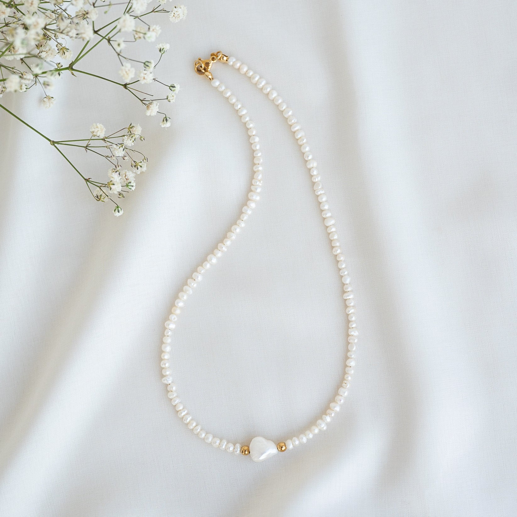 Eine zarte Perlenkette mit kleinen, gleichmäßig verteilten Perlen und einer größeren Perle in der Mitte, die von goldenen Perlen flankiert wird, auf einem weißen Stoffhintergrund mit Babyatemblüten in der Ecke.