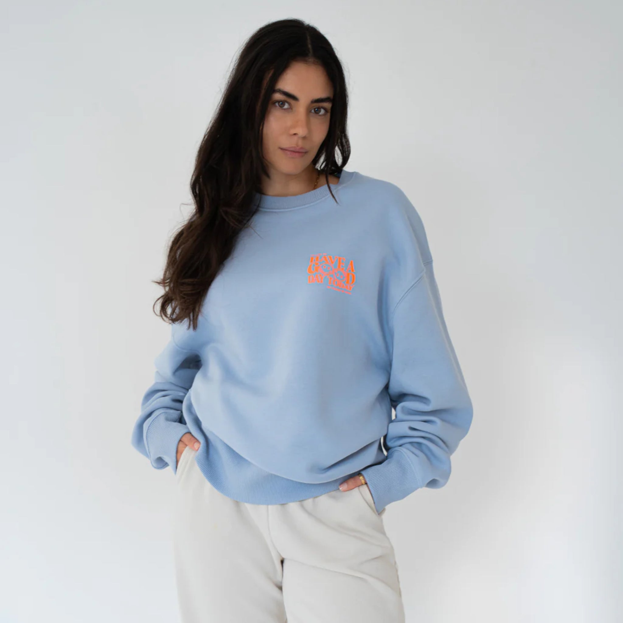 Eine Frau mit langen dunklen Haaren trägt ein hellblaues Sweatshirt und eine helle Hose. Sie steht vor einem schlichten weißen Hintergrund, hat die Hände in den Taschen und schaut ruhig in die Kamera.