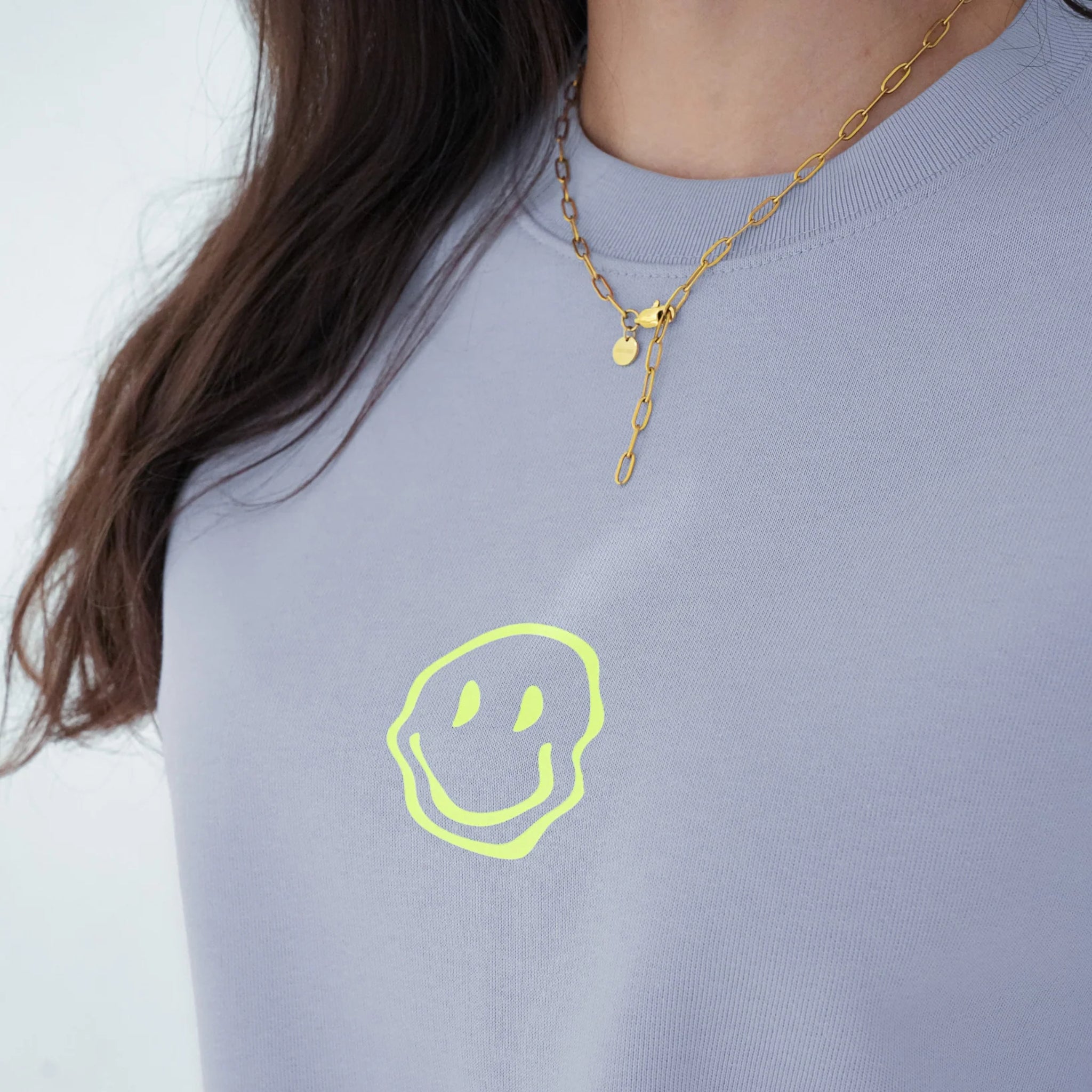 Eine Person, die eine goldene Halskette und ein hellviolettes Hemd mit einem neongelben, handgezeichneten Smiley auf der Brust trägt.
