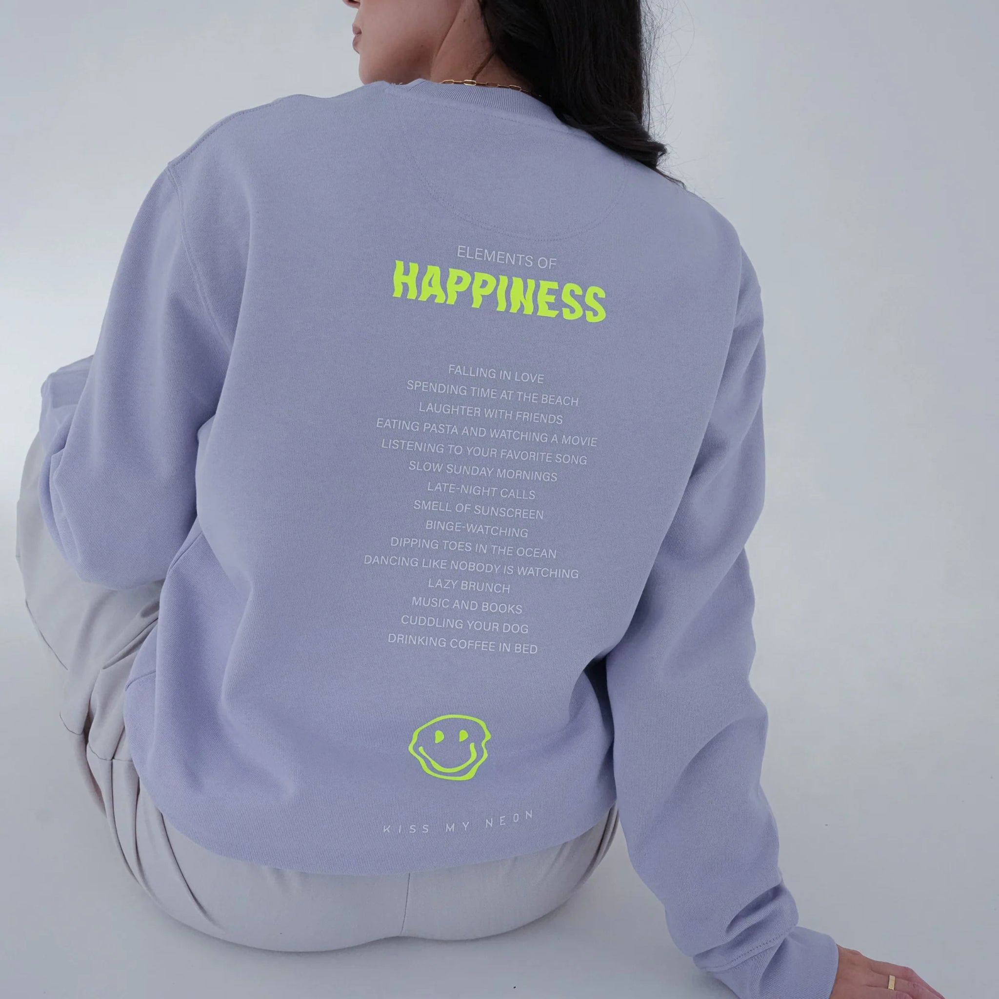 Eine Frau sitzt mit dem Rücken zur Kamera und trägt ein hellviolettes Sweatshirt mit gelber Aufschrift "Elements of Happiness" und einer Smiley-Grafik am unteren Saum.