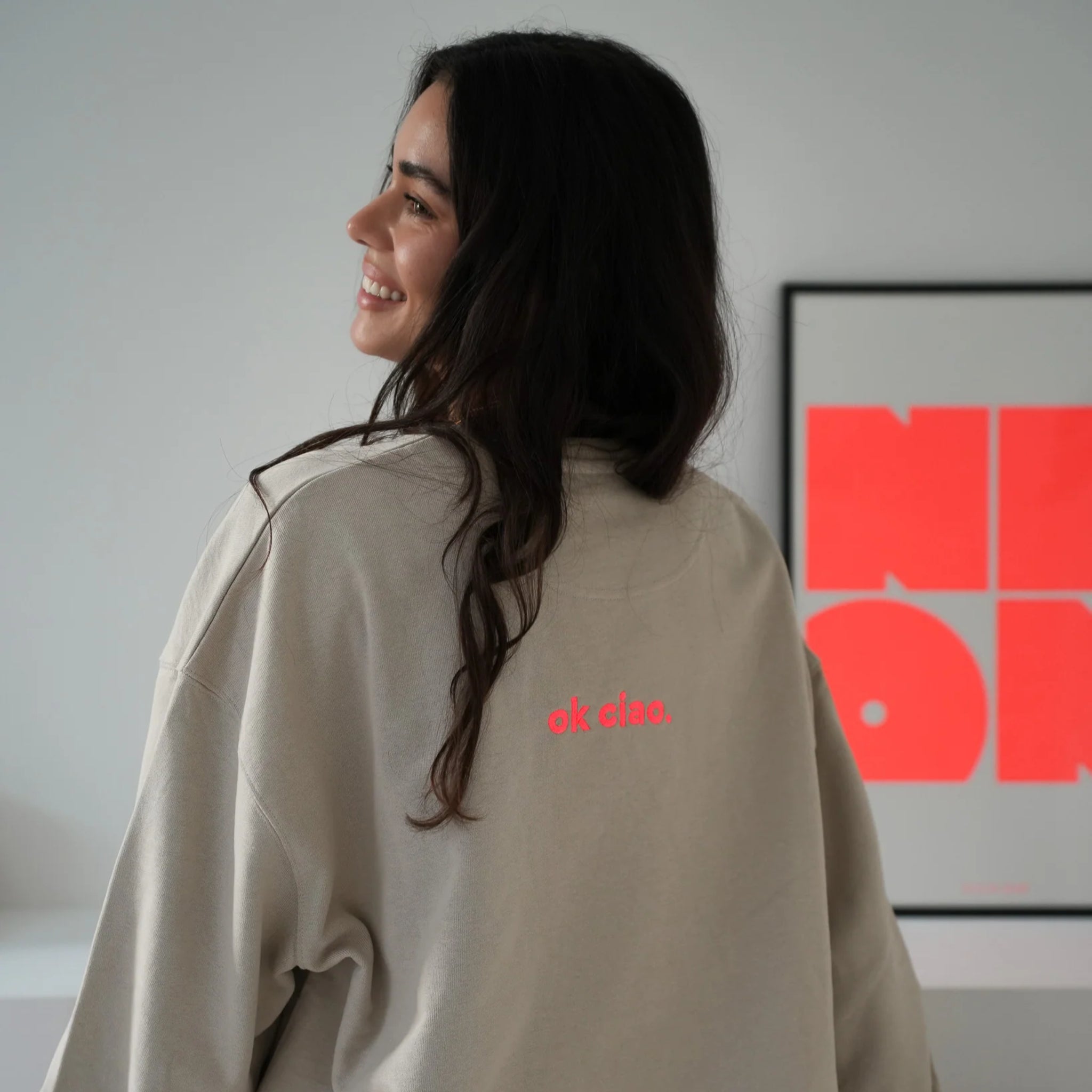 Eine Frau mit langen dunklen Haaren lächelt, als sie über ihre Schulter blickt. Sie trägt ein beiges Sweatshirt mit der Aufschrift ok ciao. in rosa auf dem Rücken. Hinter ihr hängt ein moderner Kunstdruck mit fetten roten Buchstaben an der Wand.