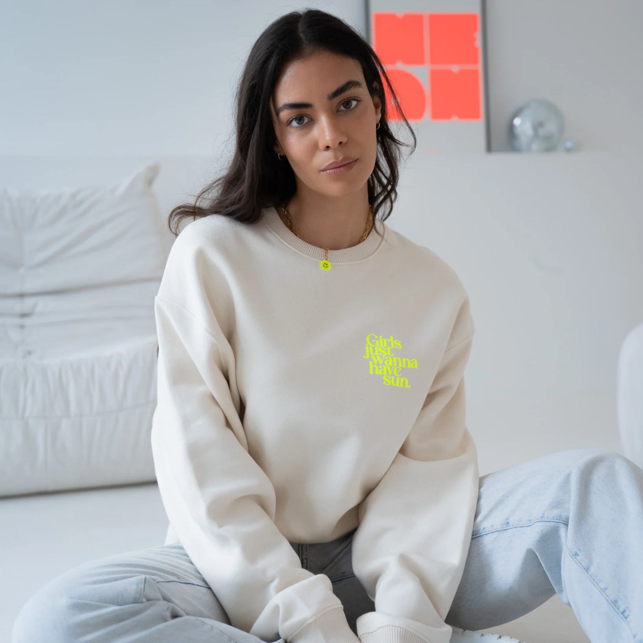Eine Frau mit langen dunklen Haaren sitzt in einem Haus auf dem Boden. Sie trägt ein cremefarbenes Sweatshirt mit gelber Aufschrift und hellblaue Jeans. Sie blickt mit neutralem Blick in die Kamera. Der Hintergrund ist minimalistisch und weiß.