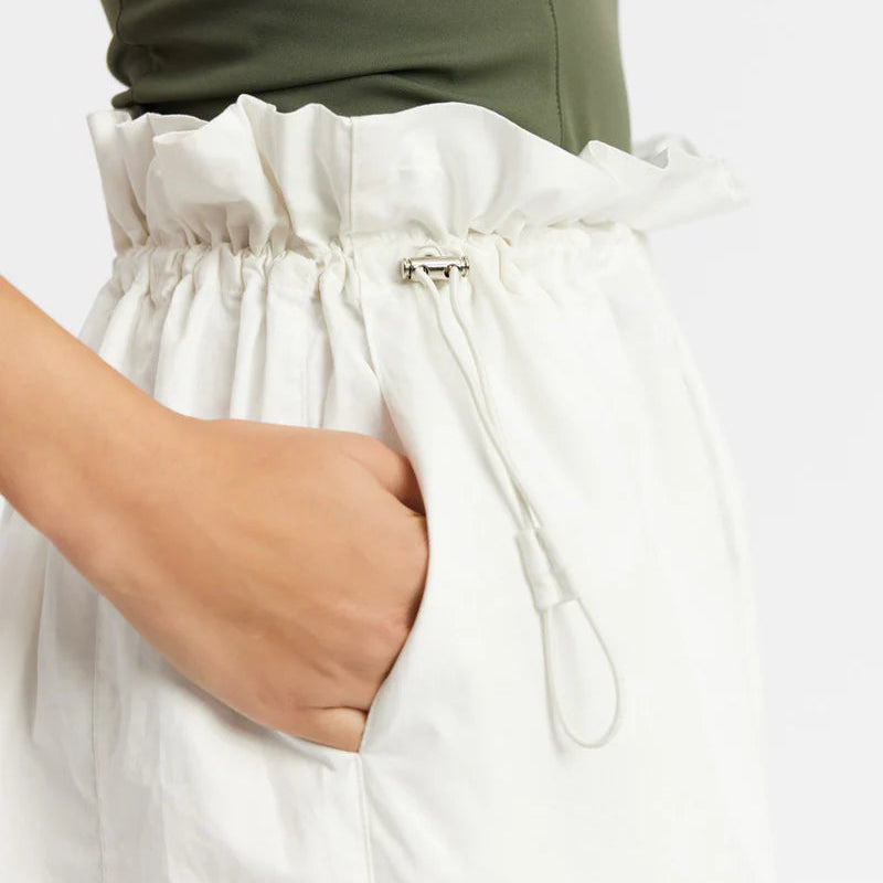 Eine Person, die ein ärmelloses olivgrünes Oberteil und eine hochgeschnittene weiße Papiertütenhose mit einem verstellbaren Kordelzug trägt, wird mit einer Hand in einer Tasche vor einem unifarbenen Hintergrund gezeigt.