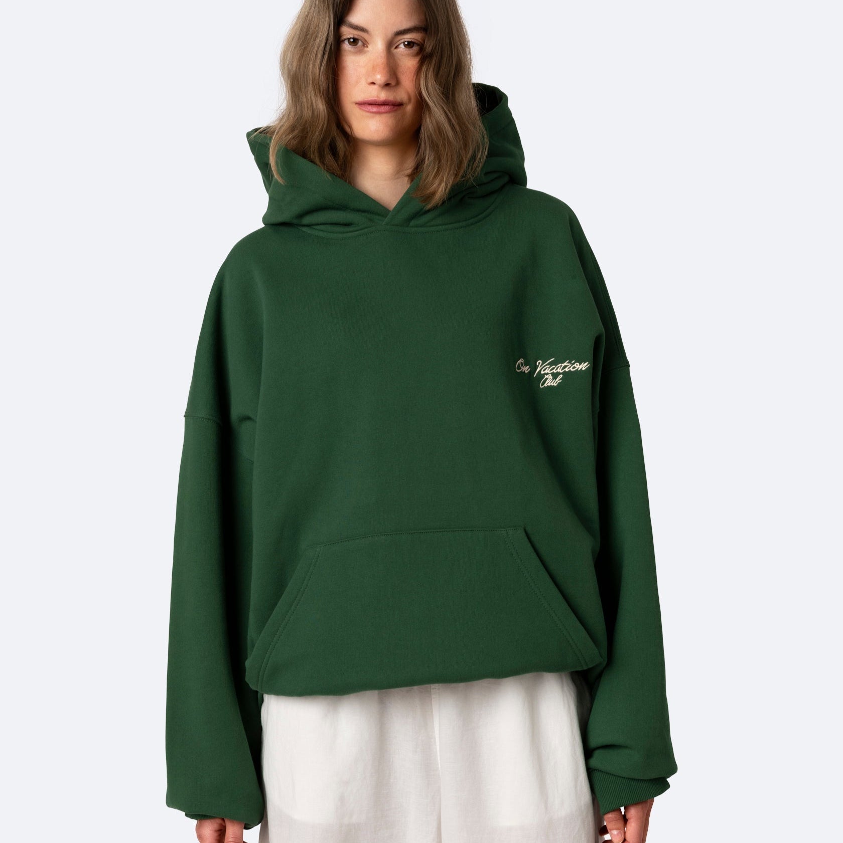 Eine Person, die ein dunkelgrünes Kapuzensweatshirt mit weißer Aufschrift auf der Brust und einen weißen Rock trägt, steht vor einem schlichten hellen Hintergrund.