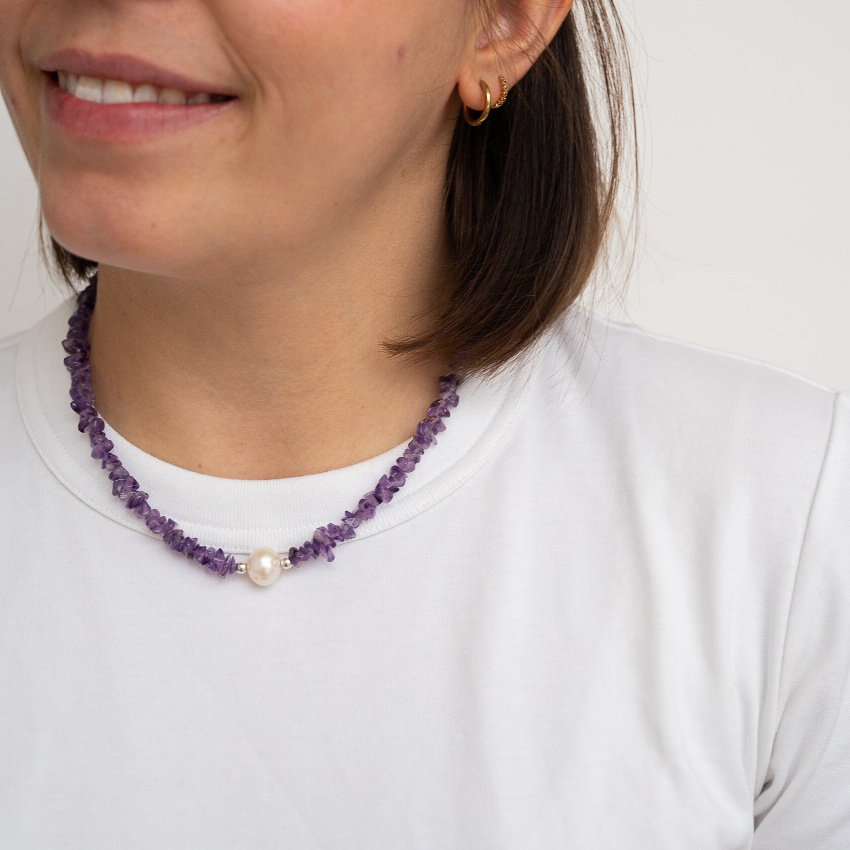 Eine Frau trägt ein weißes T-Shirt und eine lila Perlenkette mit einer einzigen großen Perle in der Mitte. Sie hat kurzes braunes Haar und lächelt leicht, wobei ein kleiner goldener Ohrring zu sehen ist.