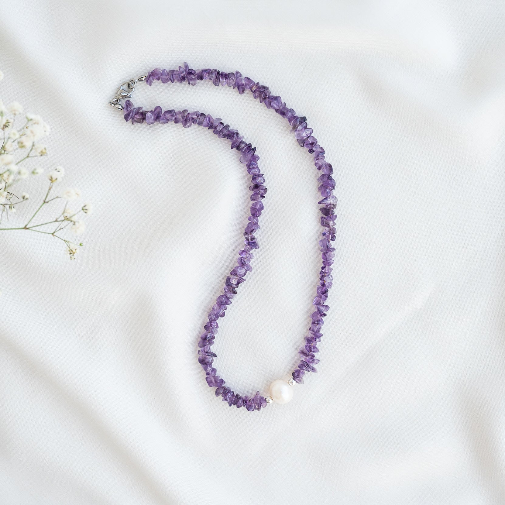 Eine Halskette aus unregelmäßigen violetten Edelsteinperlen mit einer einzelnen großen weißen Perle in der Mitte, die auf einem weißen Stoffhintergrund mit kleinen weißen Blumen in der Ecke angebracht ist.