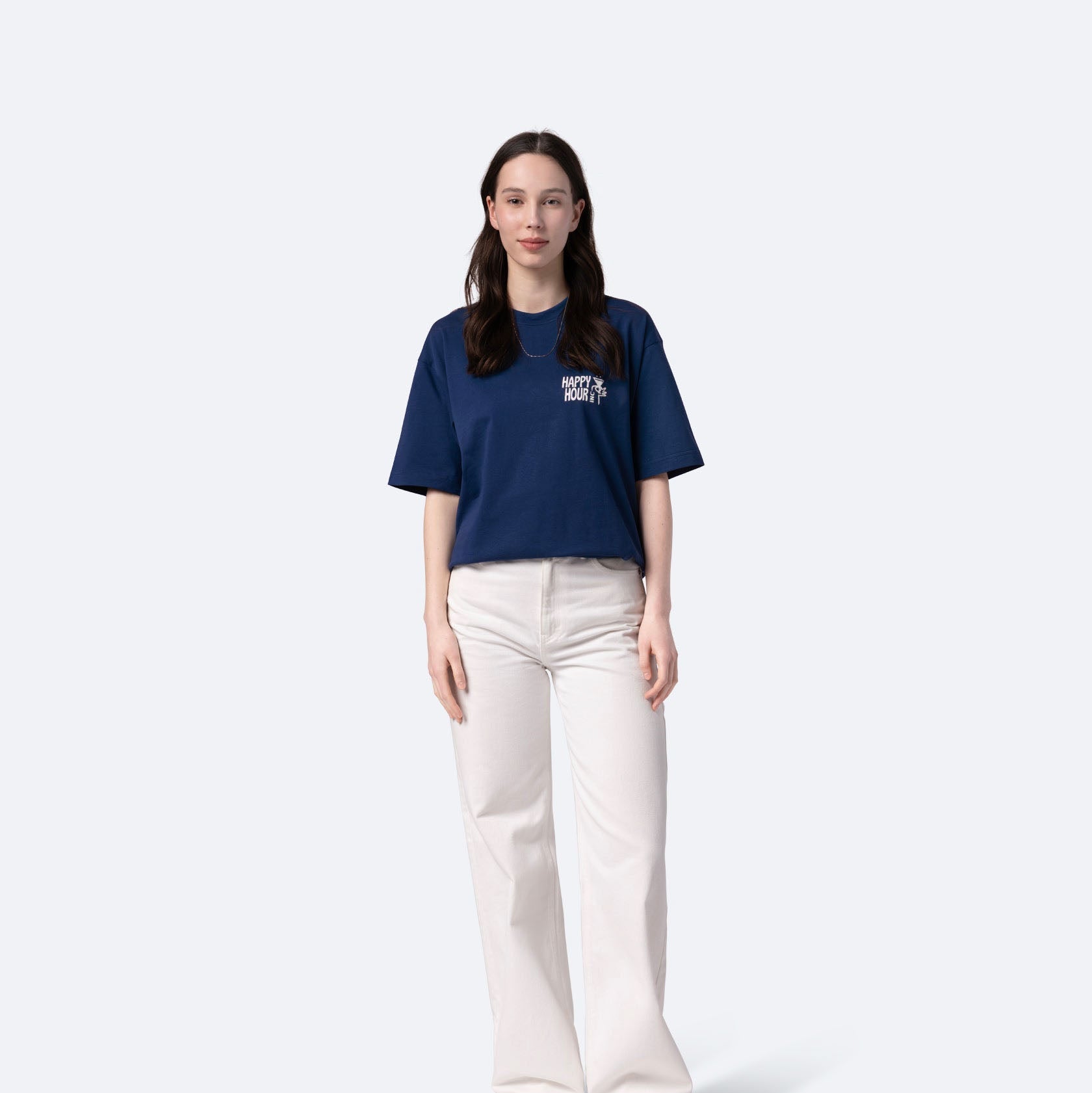 Eine Frau mit langen dunklen Haaren steht mit dem Gesicht nach vorne, trägt ein marineblaues T-Shirt, weiße Hosen und weiße Schuhe vor einem schlichten hellen Hintergrund.