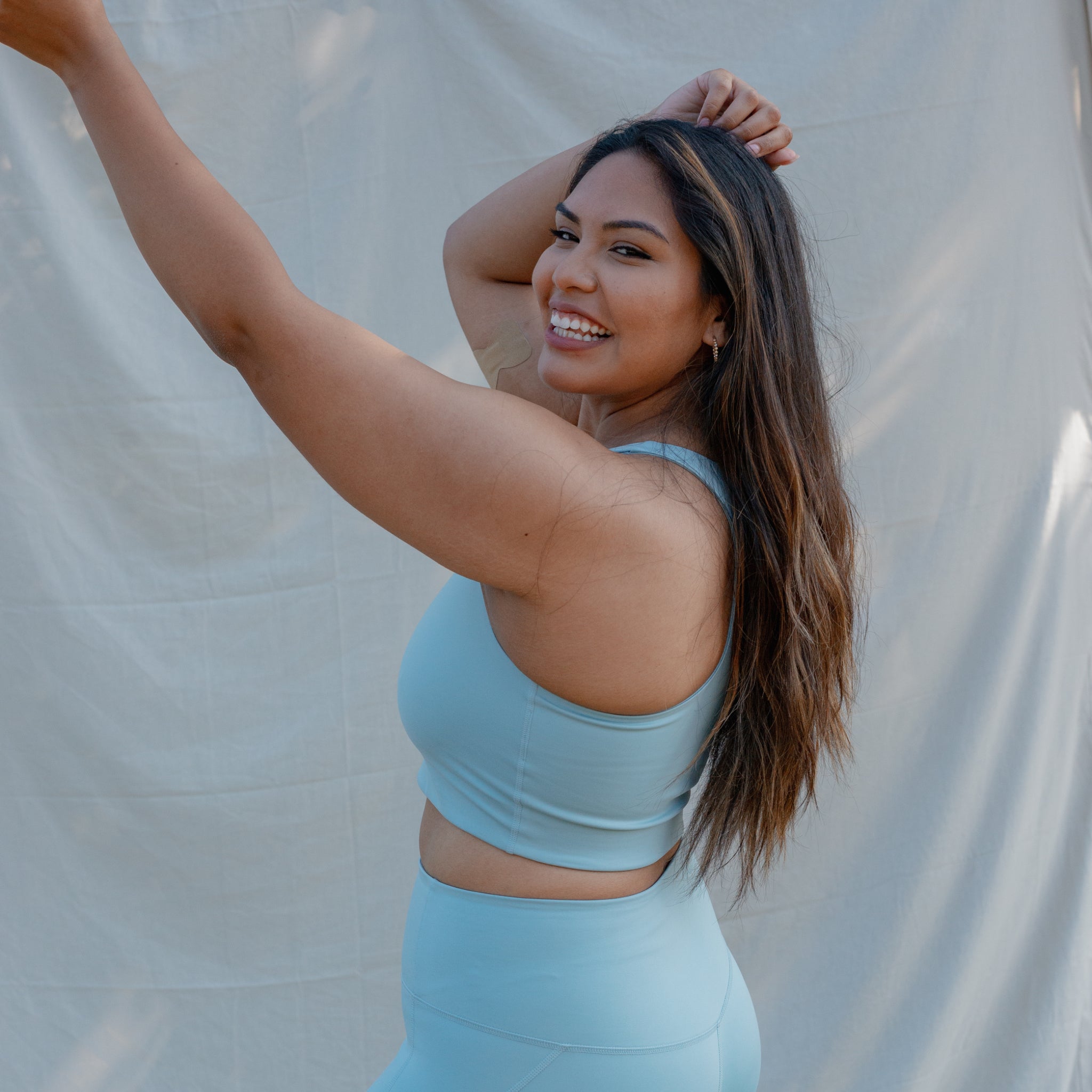 Eine Frau in hellblauer Sportkleidung lächelt und hebt die Arme vor einem beigen Hintergrund.