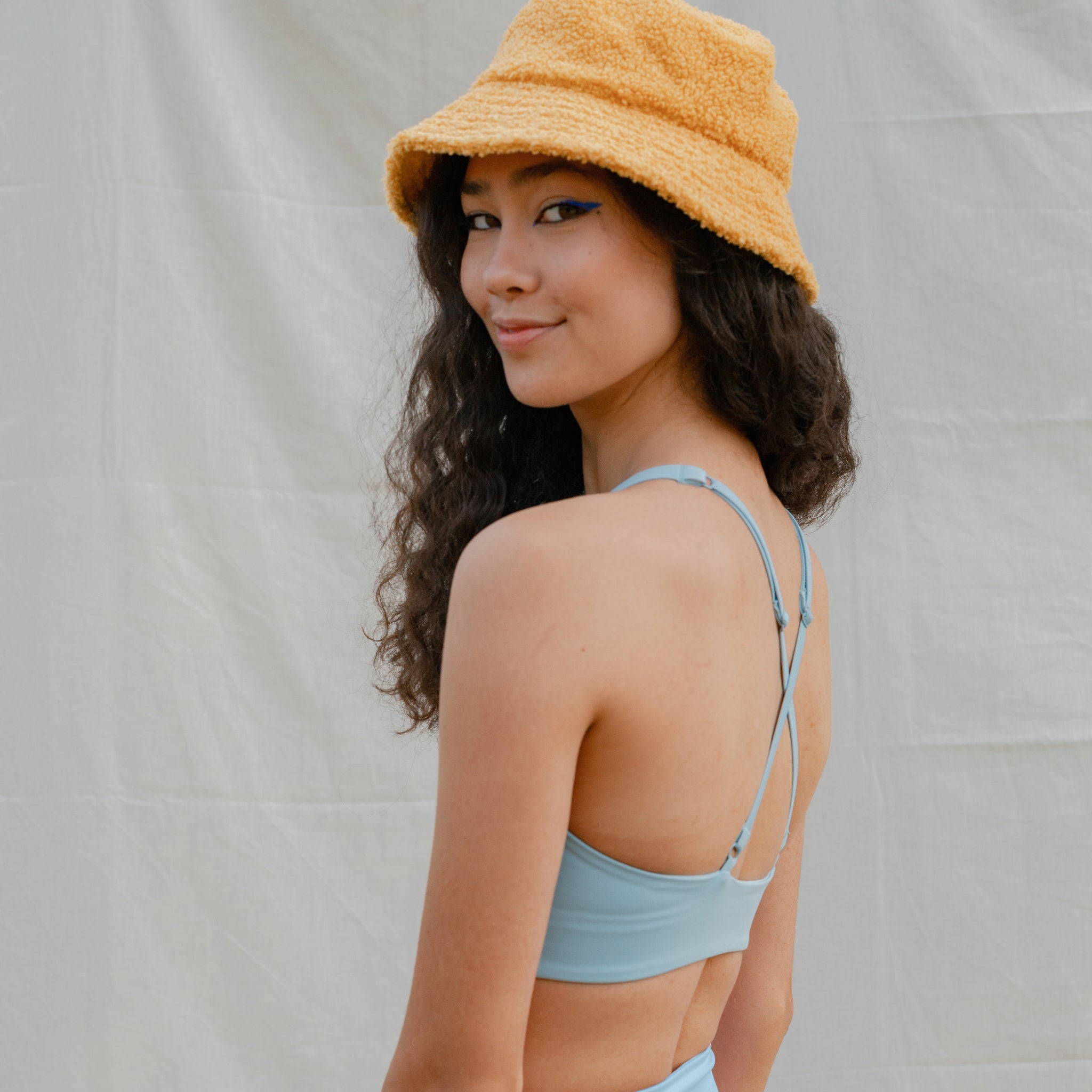 Eine Frau mit gelbem Hut und blauem Oberteil blickt über die Schulter und lächelt vor einem neutralen Hintergrund.