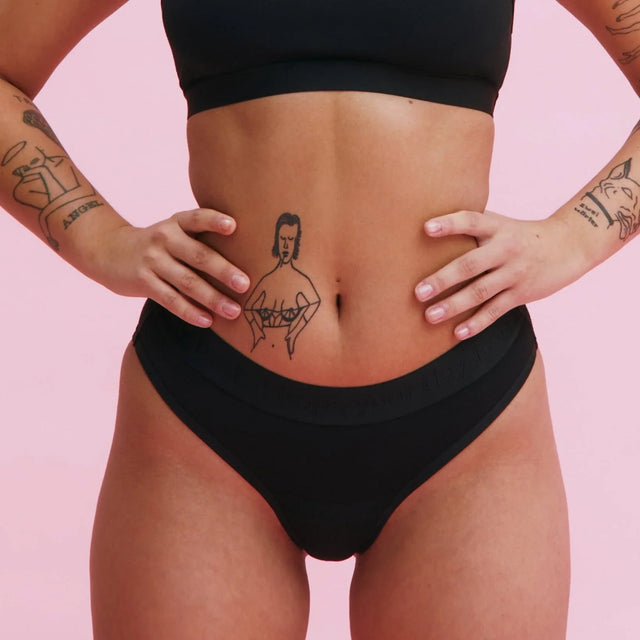 Eine Person, die schwarze Unterwäsche und ein schwarzes Oberteil trägt, steht mit den Händen in den Hüften vor einem rosa Hintergrund. Sichtbare Tätowierungen auf den Armen und eine Tätowierung einer Frau in einem BH auf dem Unterbauch.