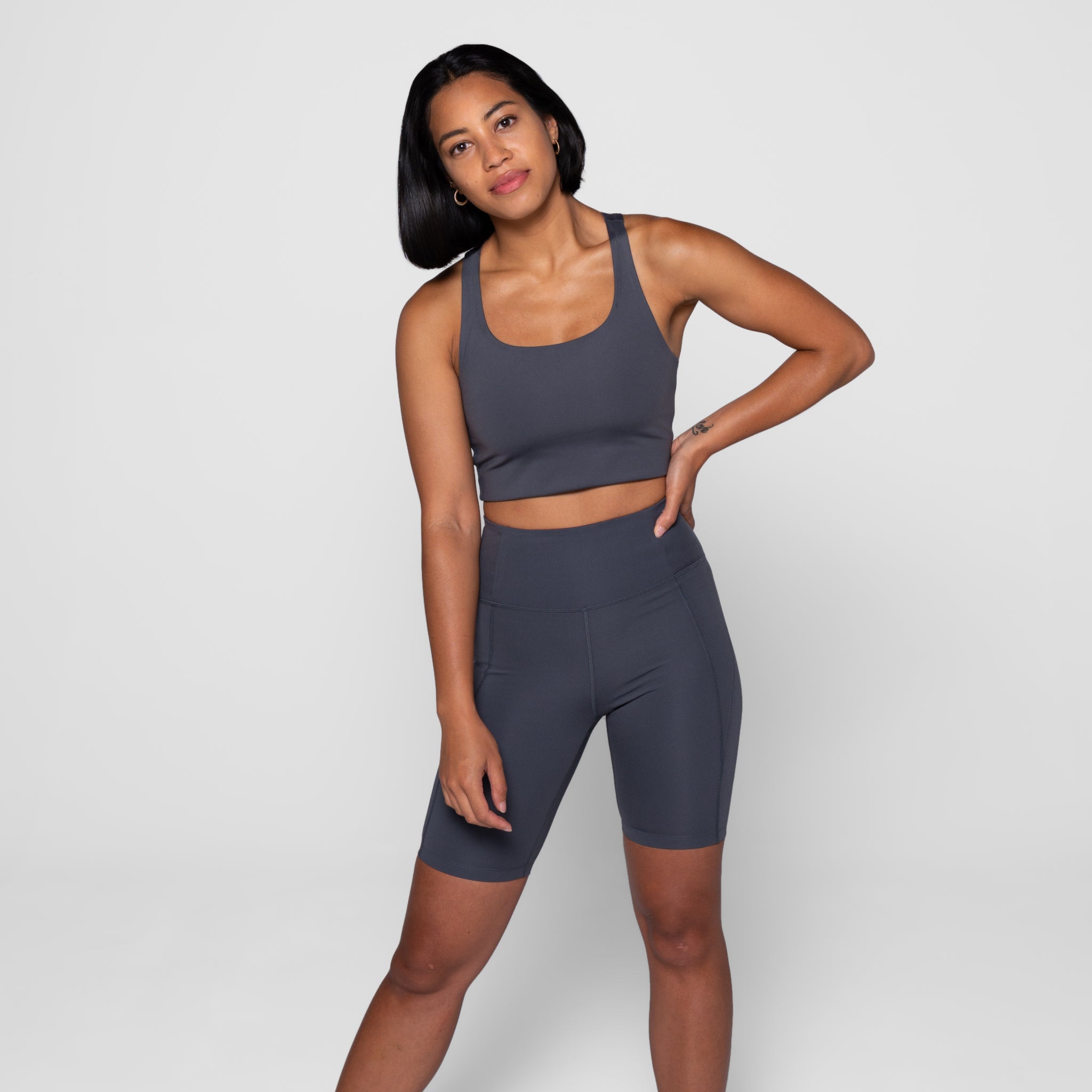 Eine Frau in einem grauen Sporttop und -shorts steht vor einem neutralen Hintergrund.