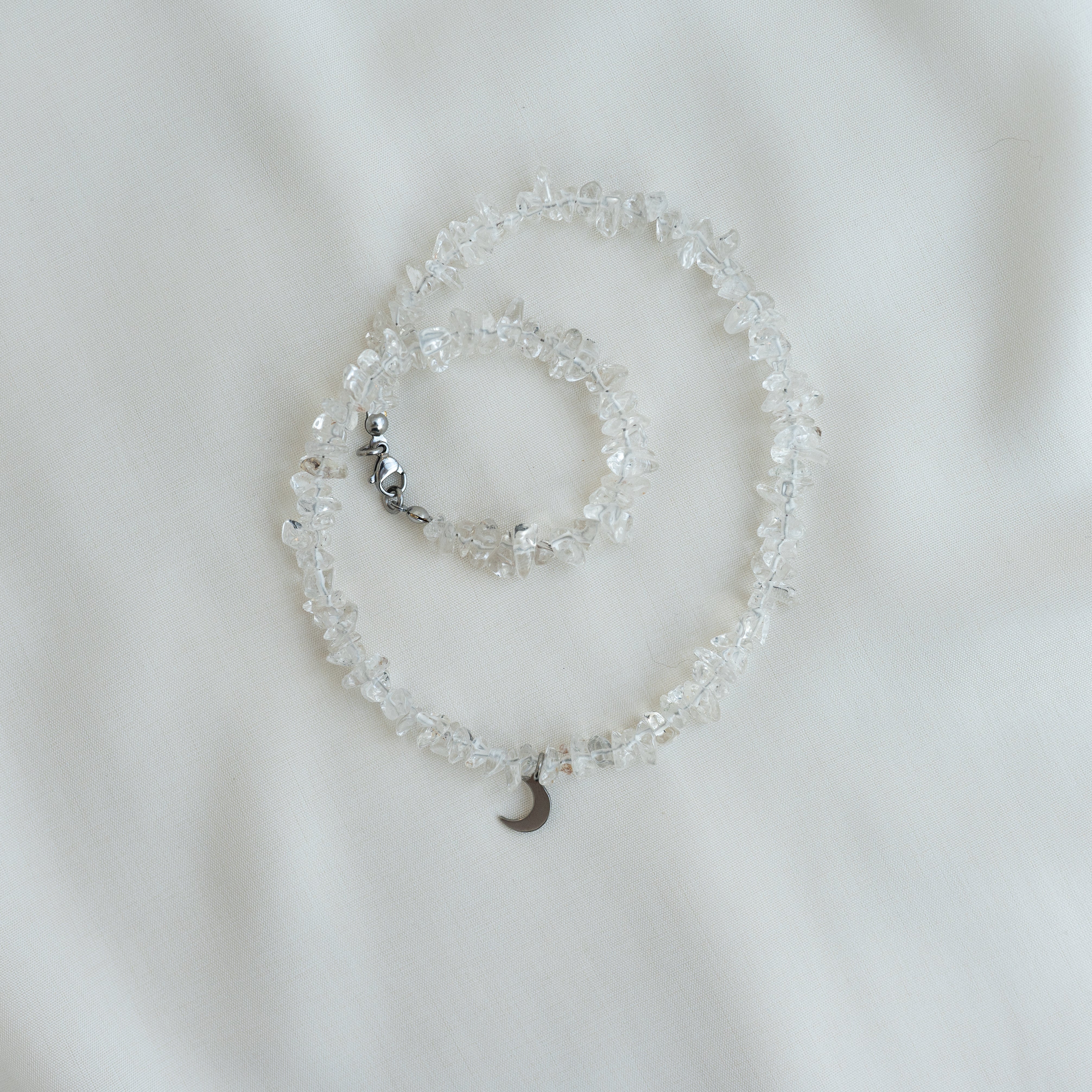 Eine Kette mit Chips in klarem Kristall, einem kleinen Mondsichel-Charme und einem silbernen Verschluss ist spiralförmig auf einem weichen, weißen Stoffhintergrund angeordnet.