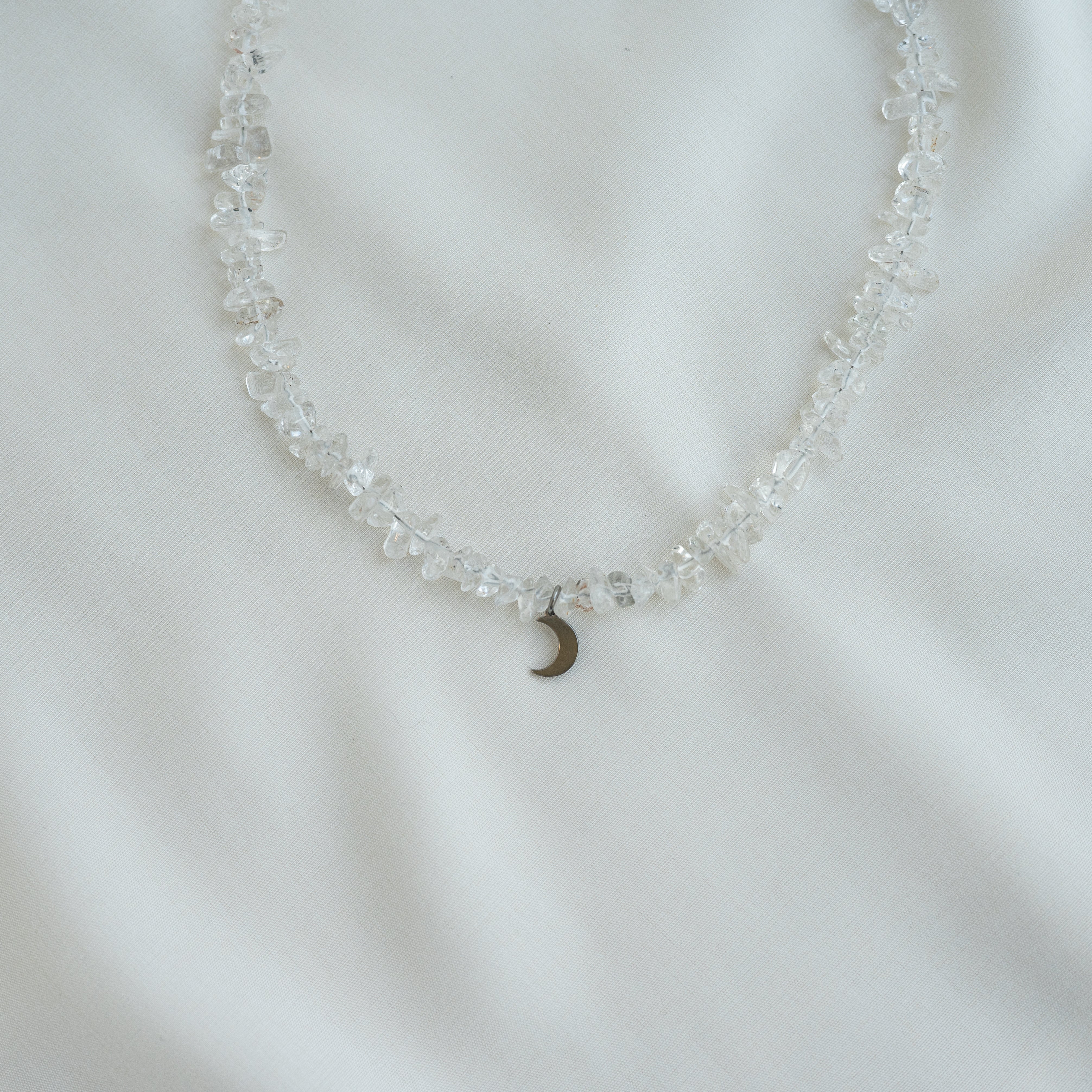 Eine zarte Halskette aus klaren, unregelmäßig geformten Perlen mit einem kleinen Halbmond-Anhänger liegt auf einem weißen Stoffhintergrund.