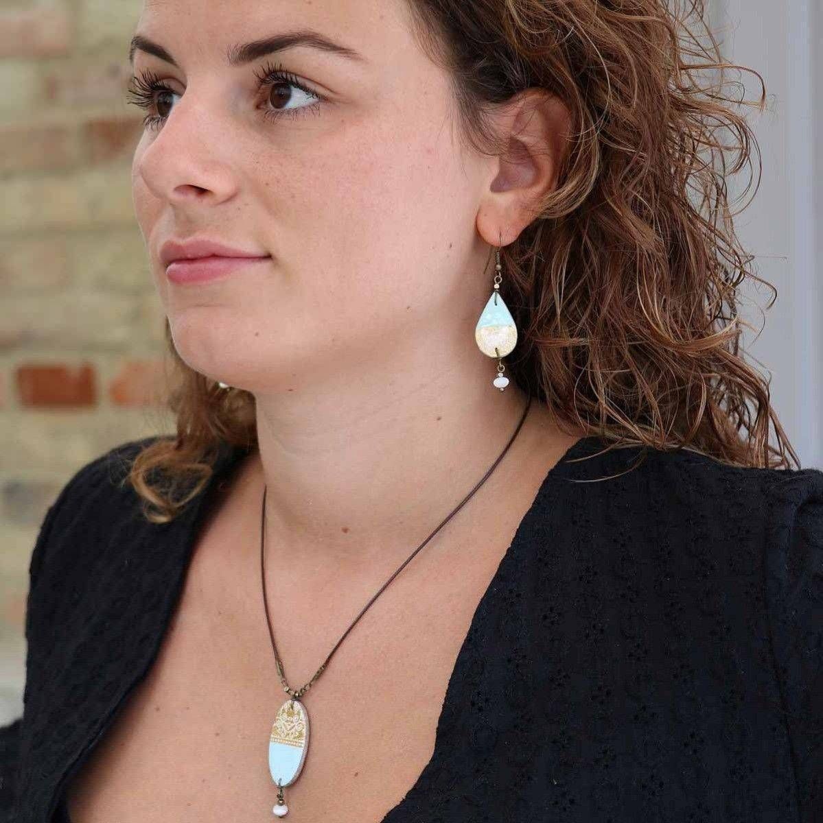 Eine Frau mit lockigem braunem Haar trägt ein schwarzes strukturiertes Oberteil und ein dazu passendes Schmuckset mit einem tropfenförmigen Anhänger an der Halskette und Ohrringen mit weißen und goldenen Akzenten. Sie blickt leicht zur Seite.