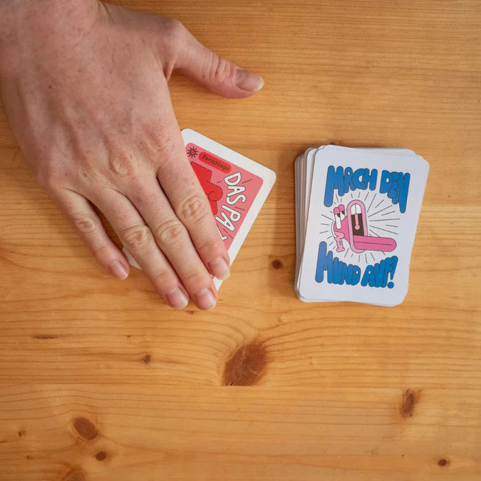 Eine Hand legt Karten auf einen Tisch, daneben liegt ein Stapel mit Karten, auf denen "Mach den Mund auf!" steht.