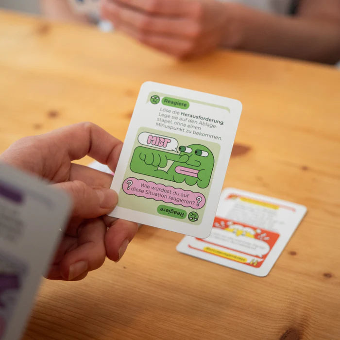 Person hält eine Spielkarte mit einer Zeichnung und Anleitung, auf einem Holztisch liegen weitere Karten.