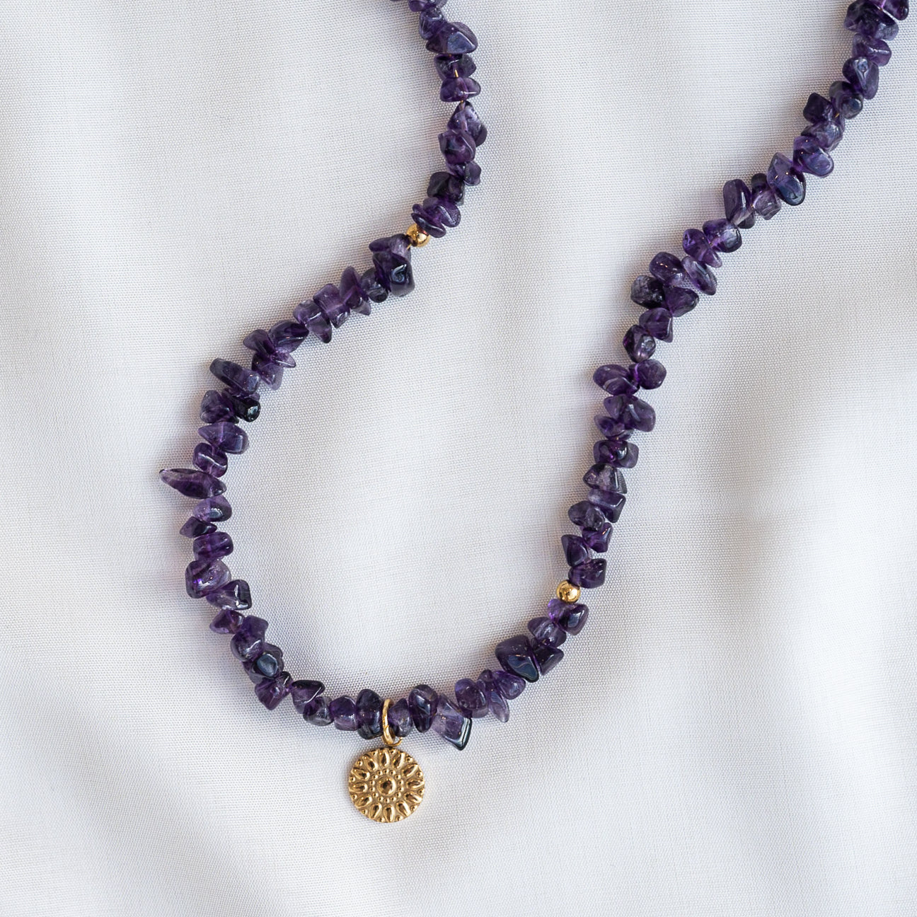 Eine Halskette aus unregelmäßigen Amethyst-Edelsteinperlen mit einem kleinen runden Goldanhänger mit Sonnenschliff-Muster auf weißem Stoffhintergrund.