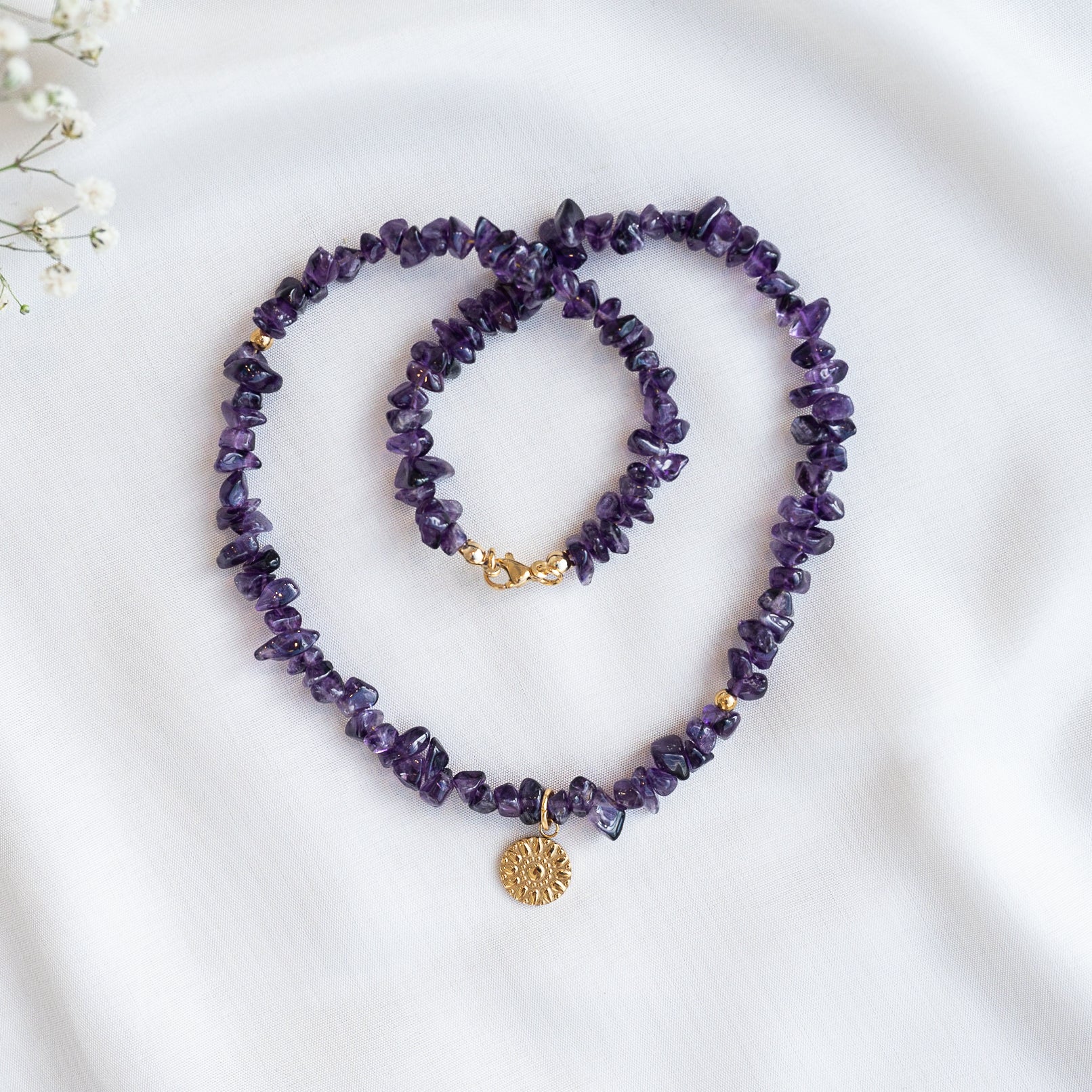 Eine Halskette und ein Armband aus unregelmäßigen Amethystperlen mit goldenen Akzenten sind auf weißem Stoff ausgestellt. Die Halskette ist mit einem strukturierten goldenen Sonnenschliff-Anhänger versehen.