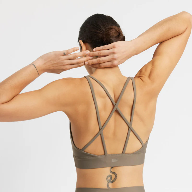 Eine Frau in einem taupefarbenen Sport-BH und Leggings steht mit dem Rücken zur Kamera, die Arme erhoben und die Hände an den Kopf gelegt. Ihr Sport-BH hat überkreuzte Träger, und sie hat ein Schlangentattoo auf dem unteren Rücken.