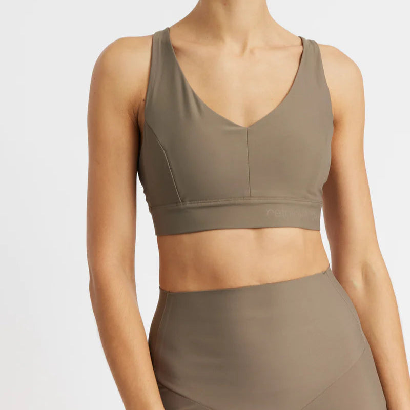 Eine Person, die einen taupefarbenen Sport-BH und dazu passende Leggings mit hoher Taille trägt, wird von den Schultern bis zur Hüfte vor einem schlichten hellen Hintergrund gezeigt.
