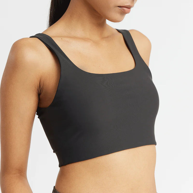 Eine Frau mit langen dunklen Haaren, die ein schwarzes, ärmelloses Crop-Top mit quadratischem Ausschnitt trägt, steht vor einem schlichten weißen Hintergrund, der von den Schultern bis zur Taille reicht.