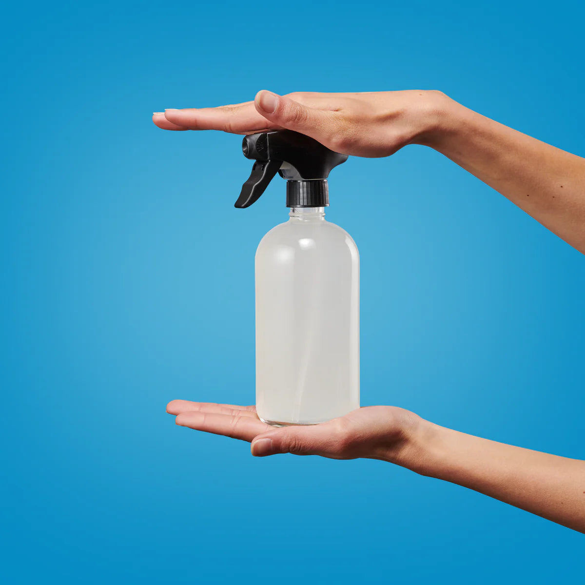 Eine Sprühflasche mit einer schwarzen Düse, die mit einer klaren Flüssigkeit gefüllt ist, wird zwischen zwei Händen vor einem soliden blauen Hintergrund gehalten. Eine Hand befindet sich unter der Flasche, die andere darüber, so dass die Flasche vertikal eingerahmt ist.