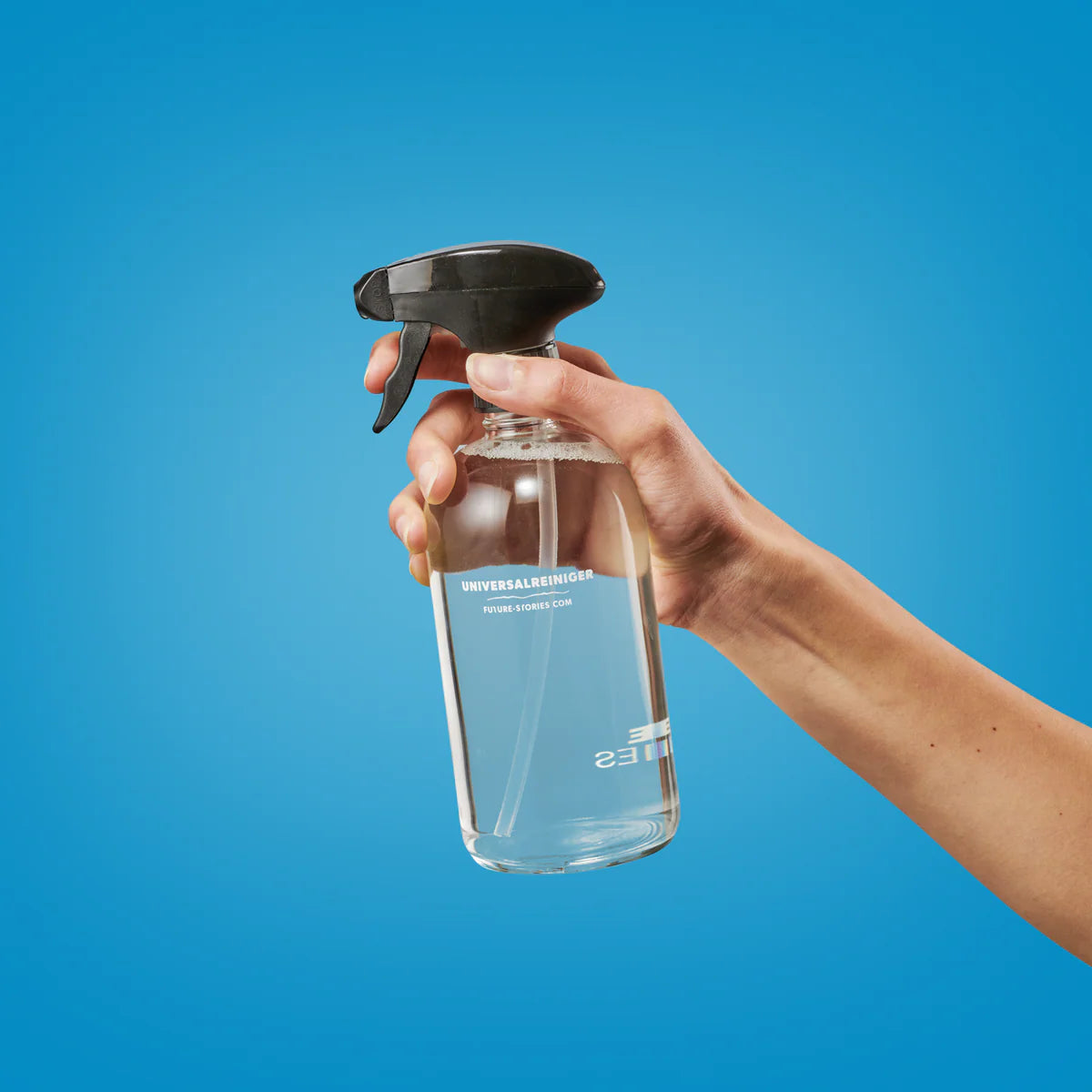 Eine Hand hält eine klare, mit Flüssigkeit gefüllte Sprühflasche vor einem einfarbig blauen Hintergrund. Die Flasche hat einen schwarzen Sprühkopf und weißen Text auf der Vorderseite.