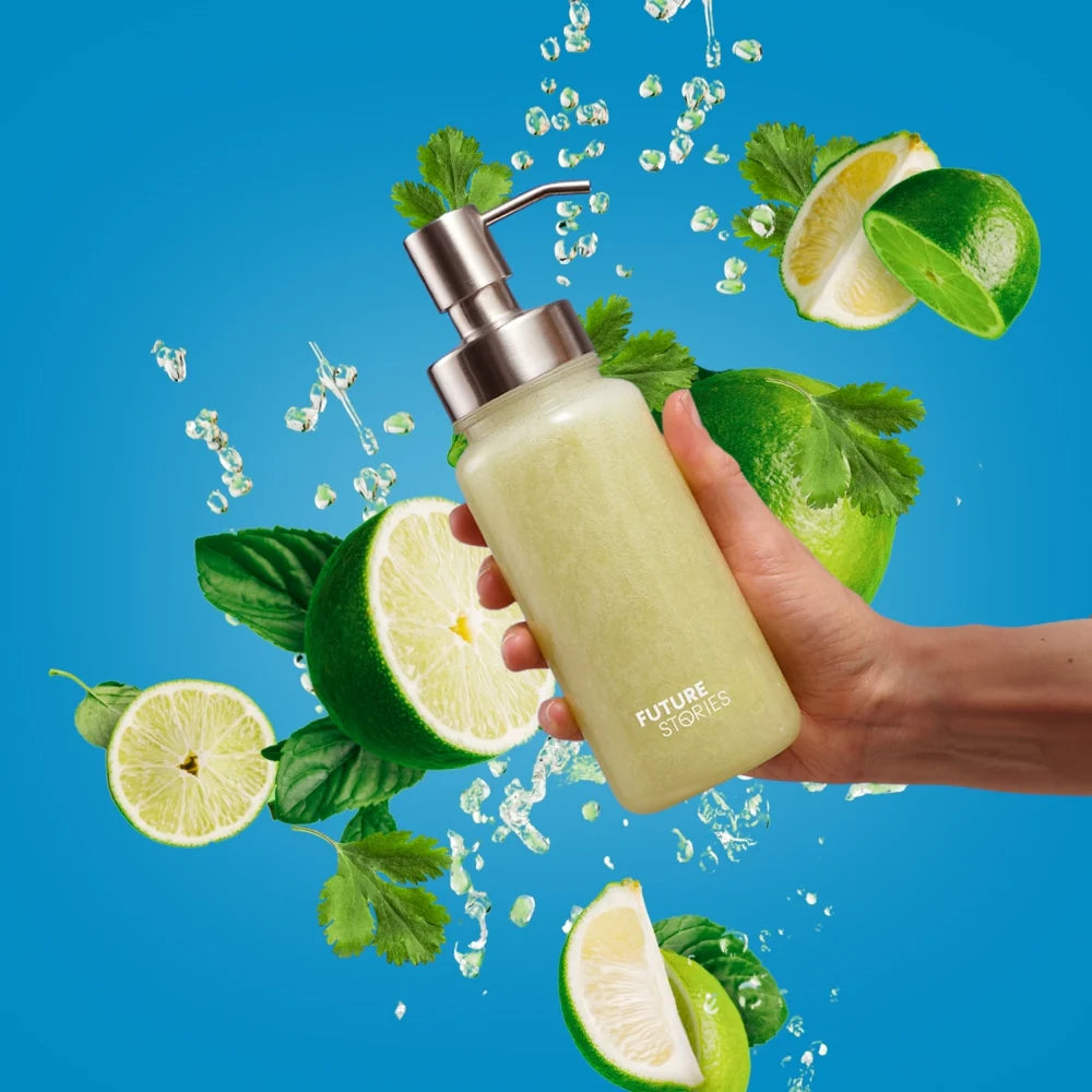 Eine Hand hält einen Seifenspender mit der Aufschrift FUTURE STORIES mit grüner Flüssigkeit, umgeben von spritzendem Wasser, Limettenhälften, Basilikum und Petersilie vor einem leuchtend blauen Hintergrund.