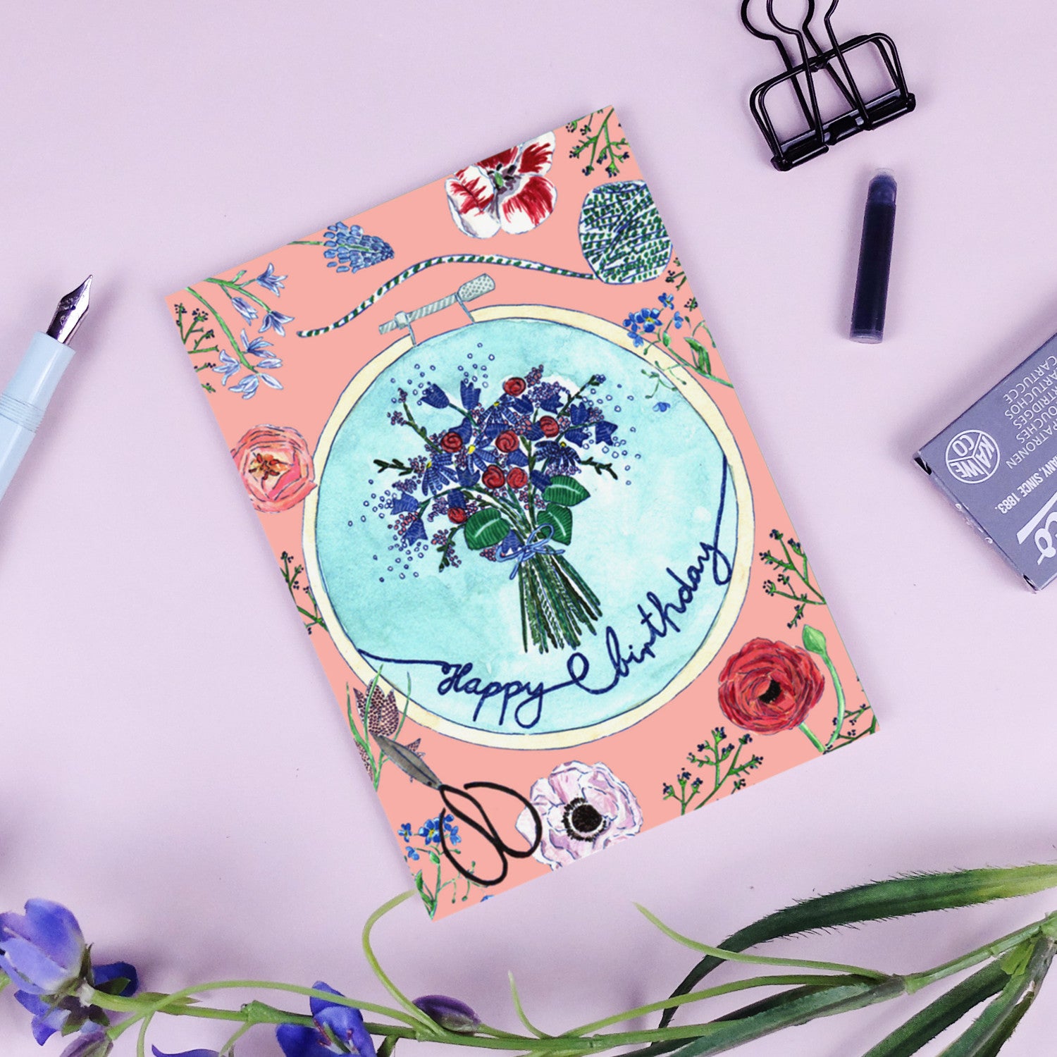 Eine Geburtstagskarte mit Blumenmotiv und der Aufschrift "Happy Birthday" liegt auf einem rosa Hintergrund mit Stift und Klammer.