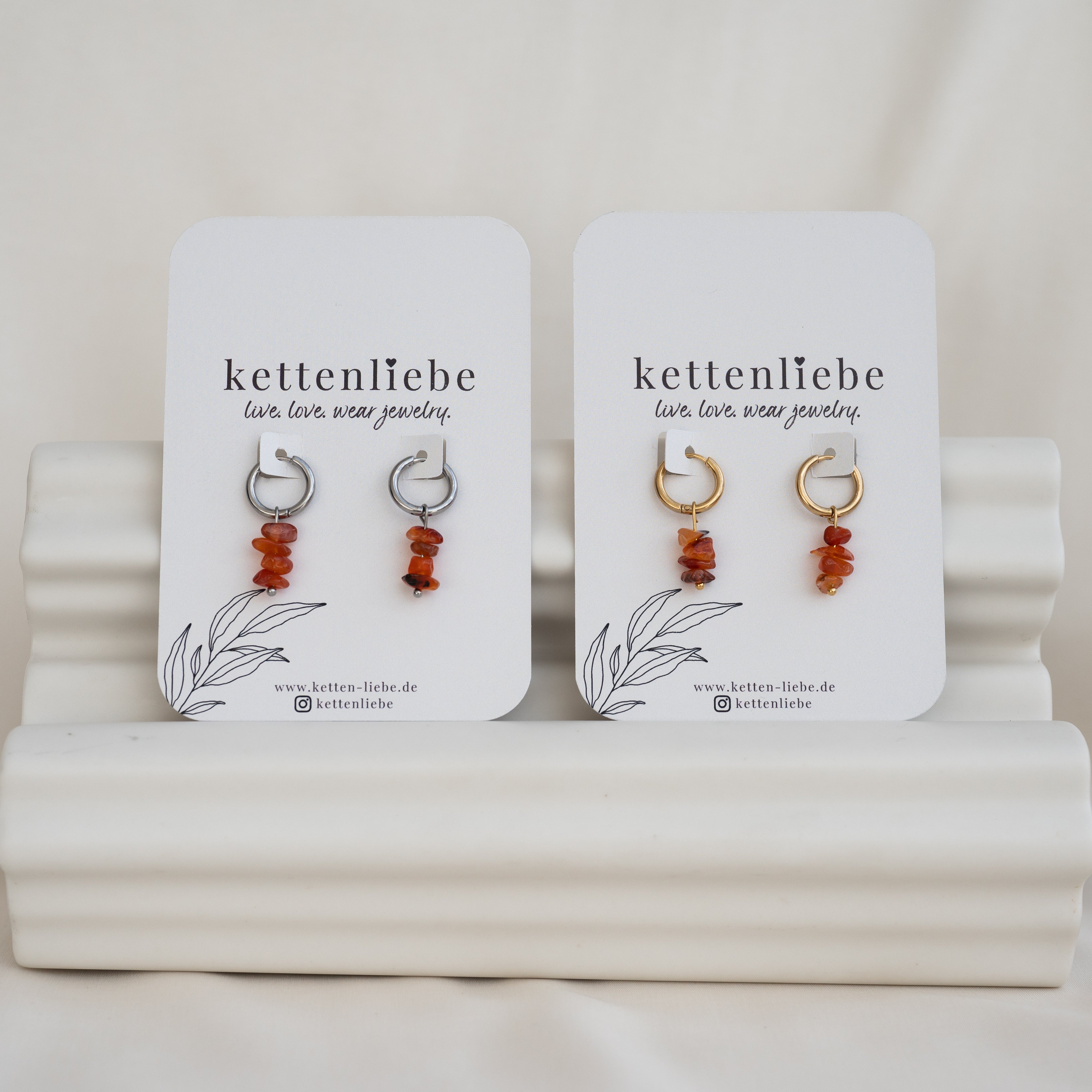 Zwei Paar Ohrringe von kettenliebe werden auf weißen Karten präsentiert. Jede Karte enthält silberne oder goldene Ohrringe mit baumelnden braunen Steinen, die auf einem weißen, gestuften Ständer befestigt sind.