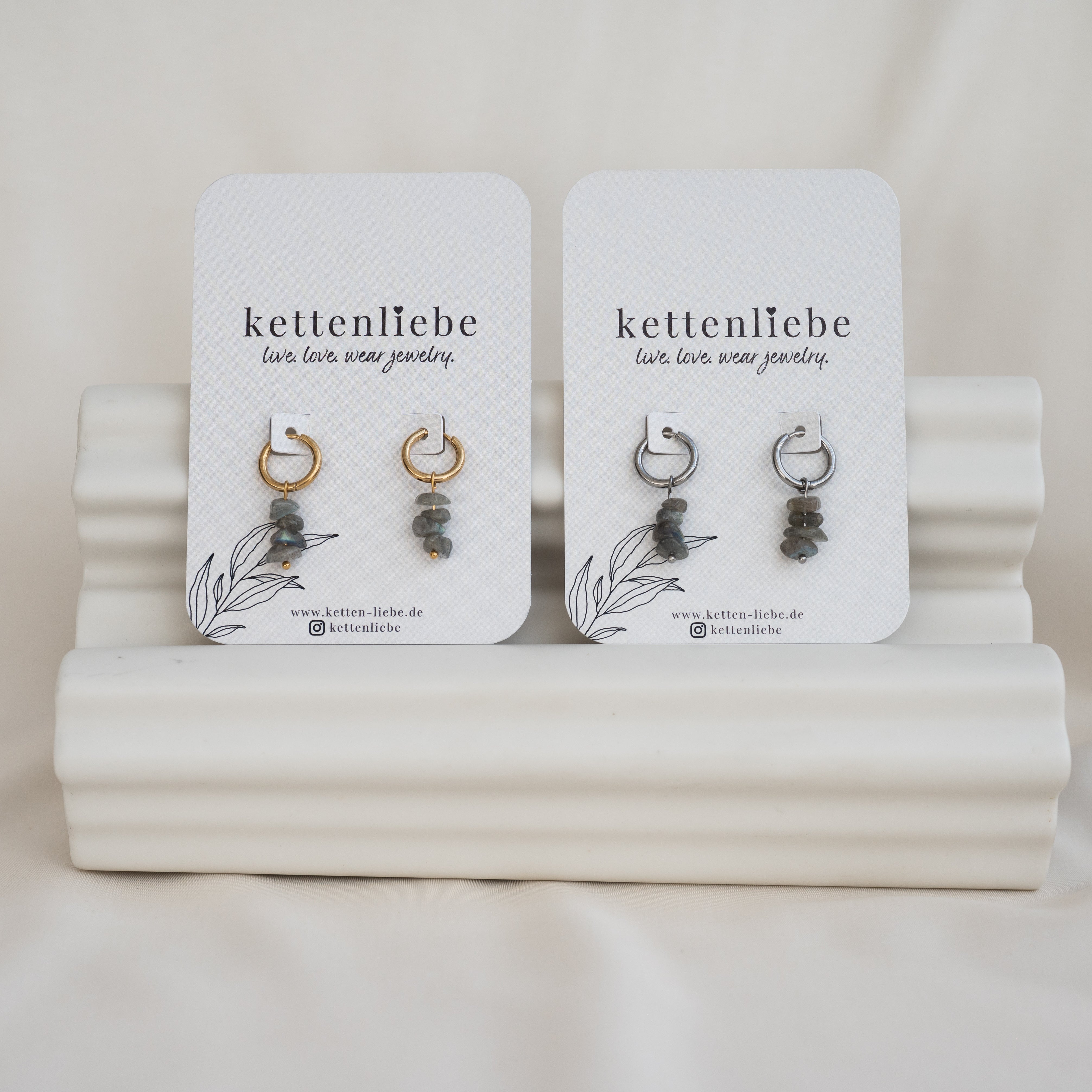 Zwei Paar Ohrhänger von kettenliebe werden auf weißen Karten mit minimalistischen botanischen Motiven präsentiert. Ein Paar hat goldene Reifen, das andere silberne, beide mit grauen Steinperlen, die auf einem strukturierten weißen Ständer stehen.