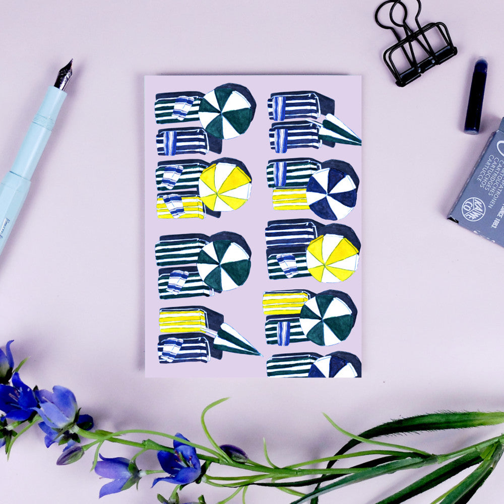 Karte mit bunten grafischen Strandmotiven, daneben ein Stift, Klammer und violette Blumen auf einem hellen Hintergrund.