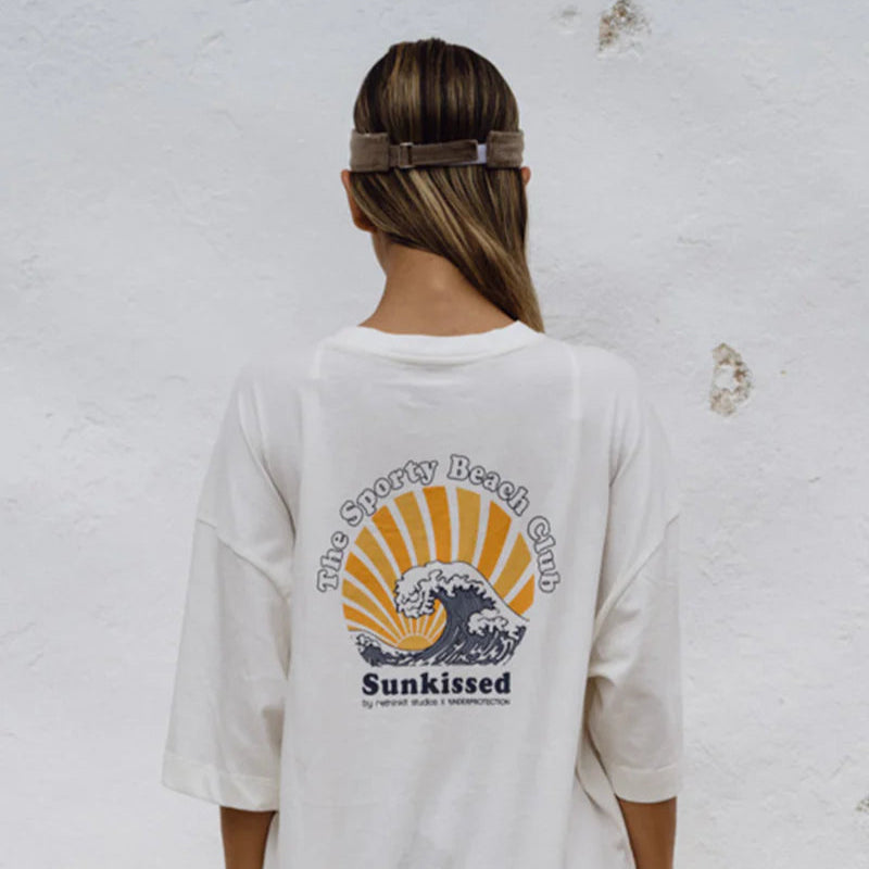 Eine Frau, die ein weißes übergroßes T-Shirt mit einer Sonnen- und Wellengrafik und der Aufschrift The Sporty Beach Club Sunkissed auf dem Rücken trägt, steht vor einer strukturierten weißen Wand. Sie trägt ein braunes Visier und hat ihr Haar zu einem tiefen Pferdeschwanz gebunden.