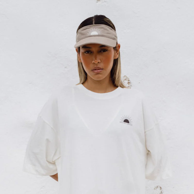 Eine Person, die einen beigen Visor und ein übergroßes weißes T-Shirt mit einer kleinen Sonnengrafik trägt, steht vor einer weißen, strukturierten Wand und schaut mit neutralem Blick direkt in die Kamera.