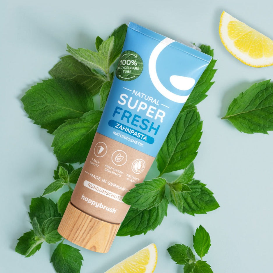 Zahnpasta-Tube "Super Fresh" von Happybrush mit Minzblättern und Zitronenscheiben auf hellblauem Hintergrund.