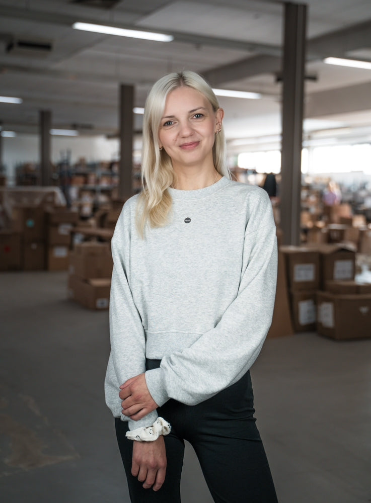 Eine lächelnde Frau steht in einem Lagerraum mit Kartons im Hintergrund, trägt einen grauen Pullover und schwarze Hose.