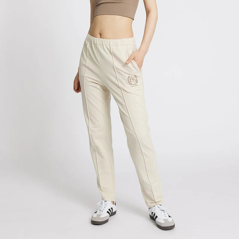 Frau in beigefarbenem Crop-Top und cremefarbener Jogginghose mit Händen in den Taschen, stehend auf weißem Hintergrund. Die Hose hat ein gesticktes Logo auf der linken Tasche, und sie trägt schwarze und weiße Turnschuhe.