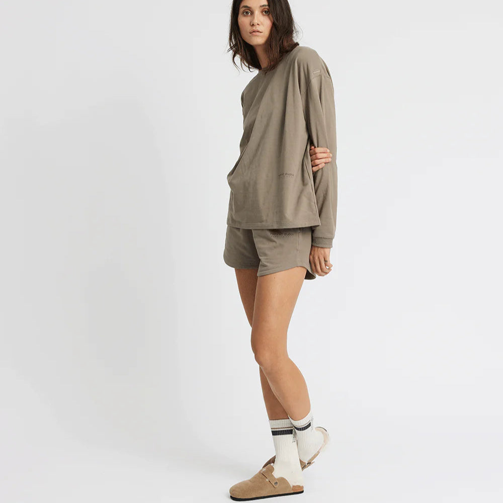 Frau in beigem Freizeitoutfit mit langen Ärmeln, Shorts und Sandalen steht vor weißem Hintergrund.