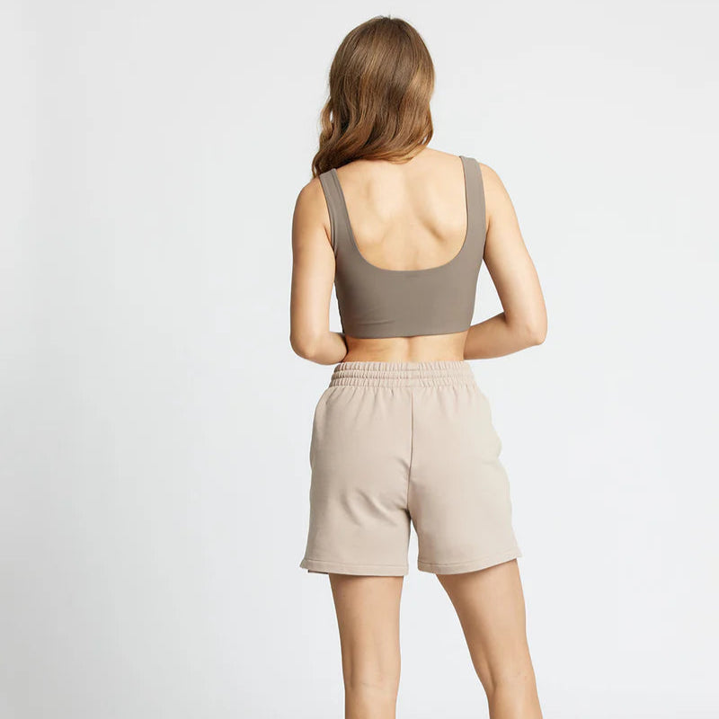 Eine Frau steht mit dem Gesicht nach hinten und trägt einen taupefarbenen Sport-BH, hellbeige Shorts und weiße Turnschuhe. Sie hat langes, hellbraunes Haar und steht vor einem schlichten weißen Hintergrund.