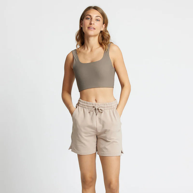 Eine Frau steht mit dem Gesicht nach vorne und trägt ein taupefarbenes Crop-Top, beigefarbene Shorts mit Kordelzug und weiße Turnschuhe. Sie hat hellbraunes Haar und einen entspannten Gesichtsausdruck, die Hände in den Taschen vor einem schlichten Hintergrund.