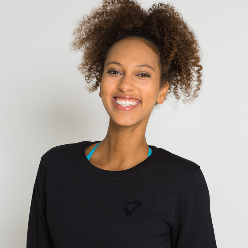 Lächelnde Person mit lockigem Haar und schwarzem Oberteil vor einem neutralen Hintergrund.