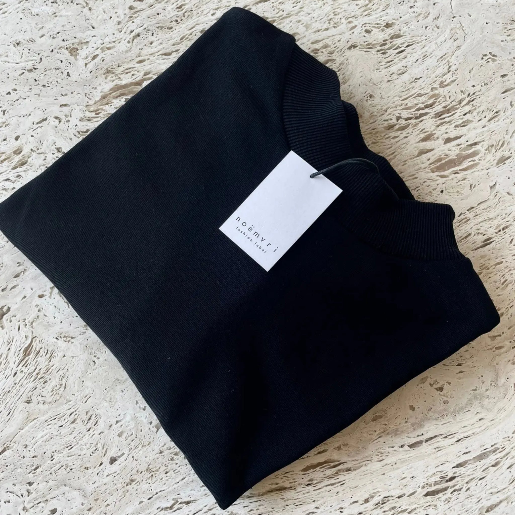 Ein fein säuberlich gefalteter schwarzer Pullover mit einem weißen Etikett auf einer hellen, strukturierten Oberfläche.