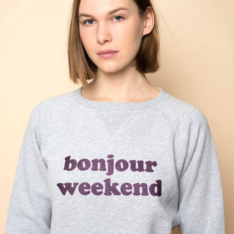 Frau mit schulterlangem Haar trägt ein graues Sweatshirt mit lila Schriftzug "bonjour weekend", vor beigem Hintergrund.
