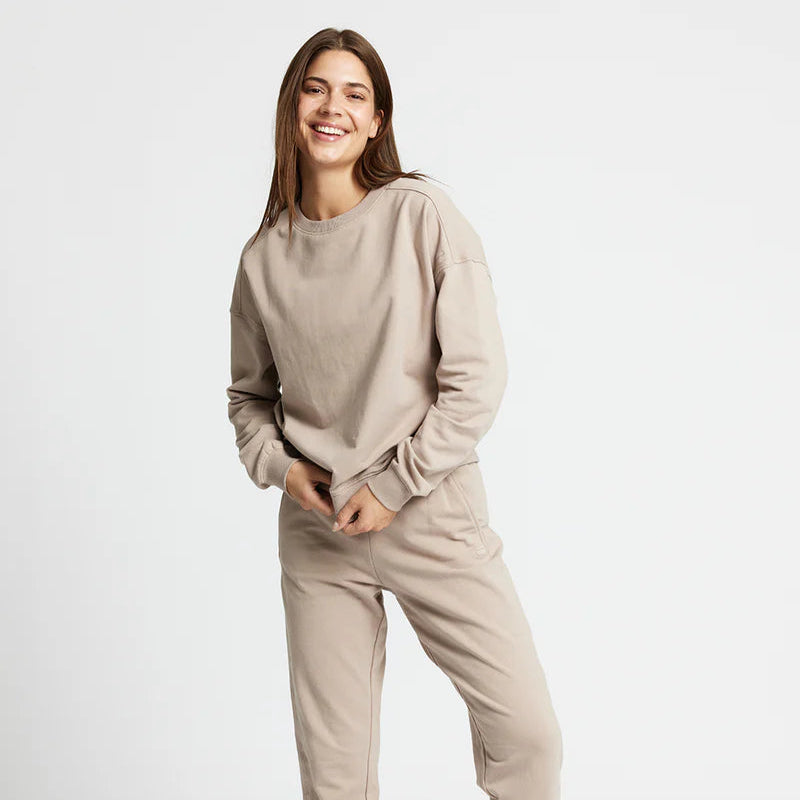 Eine Frau lächelt, während sie vor einem weißen Hintergrund steht. Sie trägt ein beiges Sweatshirt und eine passende Jogginghose mit weißen Turnschuhen.