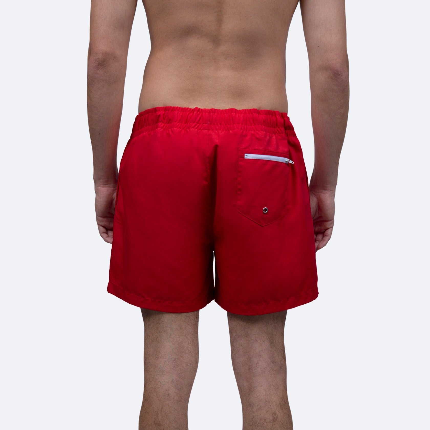 Eine Person steht von der Kamera abgewandt und trägt eine leuchtend rote Badehose mit elastischem Bund, Gesäßtasche und Reißverschluss. Die Person steht ohne Hemd vor einem schlichten, hellen Hintergrund.