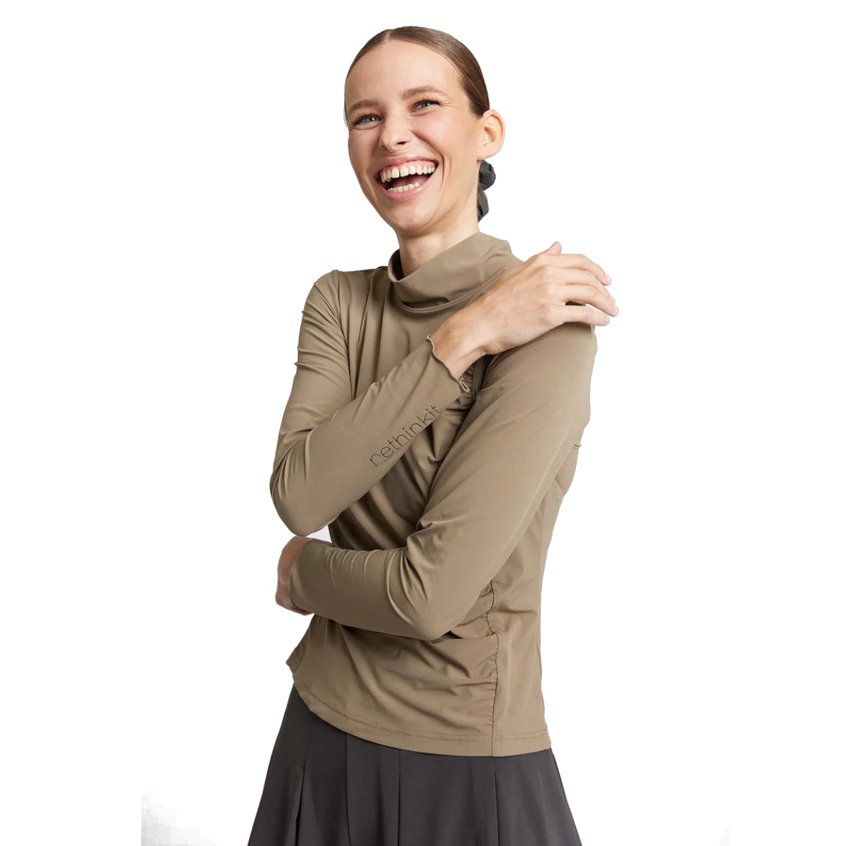 Eine lächelnde Frau mit braunem Haar in einem niedrigen Pferdeschwanz trägt ein langärmeliges, hellbraunes Rollkragenoberteil und einen dunklen Rock. Sie steht vor einem weißen Hintergrund und hat eine Hand auf ihre Schulter gelegt.