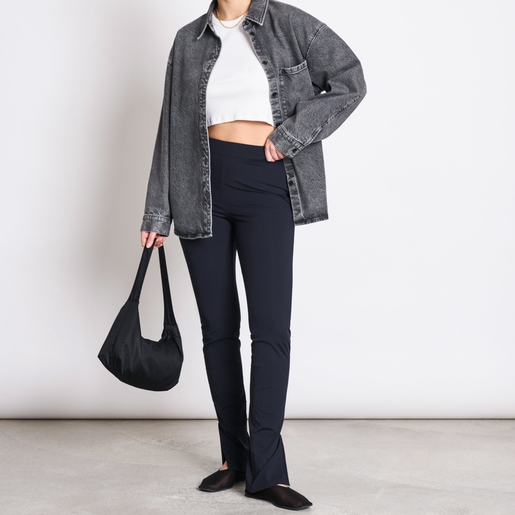 Person in grauer Jeansjacke, weißem Crop-Top und schwarzer Hose hält eine schwarze Tasche, mit neutralem Hintergrund.
